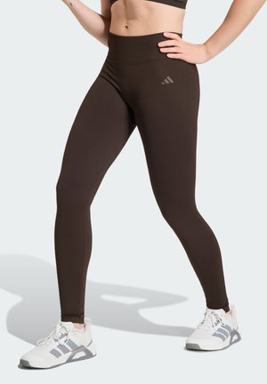 Naine, kes kannab tumepruune kõrge vöökohaga retuuse ja valgeid spordijalatseid, üks käsi puusadel, tavalise tausta ees.