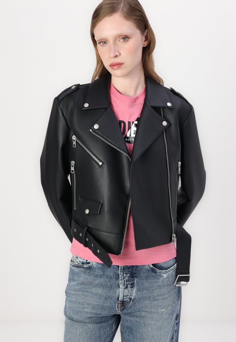Schwarze Kunstleder-Motorradjacke mit silbernen Reißverschlüssen, Druckknöpfen und Gürteldetails. Getragen über einem rosa T-Shirt und kombiniert mit blauen Jeans.