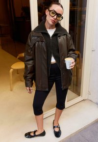 Jachetă maro din piele supradimensionată, pulover tricotat închis, leggings negri și balerini negri. Cănuță albă de cafea în mână. Ambient urban casual.