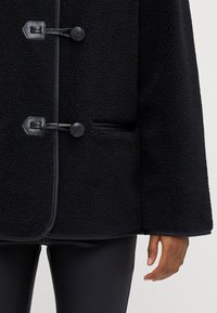 Čierny fleecový kabát s textúrovaným povrchom, zdobený čiernymi koženými zapínaniami na preklápku a bočnými vreckami, navrhnutý v uvoľnenom strihu.