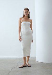 Rochie midi albă, cu o textură cu arcuri, fără bretele și cu o siluetă ajustată. Asortată cu sandale negre plate, cu barete subțiri care se încrucișează.