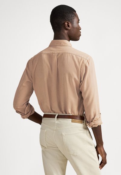 Vīrietis, skatoties prom, valkā gaiši brūnu krekls ar garām piedurknēm, kas ir sarullētas, krēmkrāsas bikses un brūnu ādas jostu, stāvot pret vienkāršu baltu fonu.