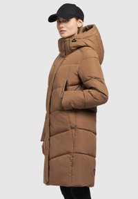Brauner Puffercoat mit Kapuze, ausgestattet mit einem gesteppte Design, Seitentaschen und einem schwarzen Logoakzent. Kombiniert mit einer schwarzen Mütze.