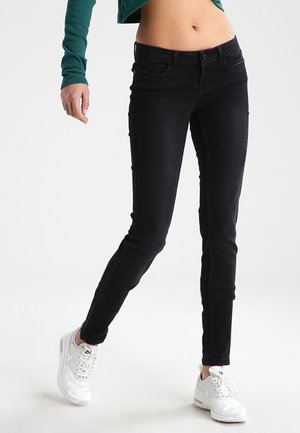 Personne portant un jean skinny noir taille haute, des baskets blanches et un top vert à manches longues court laissant voir le ventre sur un fond uni.