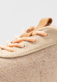 Beige Leinen-Sneaker mit orangefarbenen Schnürsenkeln, strukturiertem Obermaterial und einem braunen Lederanhänger an der Ferse. Nahaufnahme des Schnürbereichs und der Nähdetails.