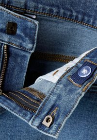 Vaqueros de denim azul con cremallera, cierre de botón y costuras en contraste. La parte interna de la cinturilla muestra una etiqueta de tela blanca y detalles de tela.