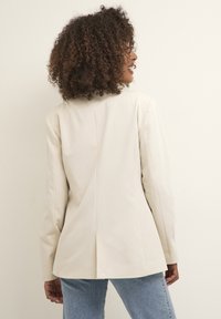 Blazer beige ajusté avec une coupe structurée, une fente unique, des manches longues et une texture lisse, associé à un jean délavé.