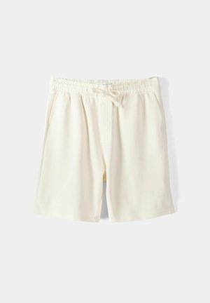Shorts de sport de couleur crème, fabriqués dans un tissu lisse, avec une ceinture élastique munie d'un cordon de serrage réglable et des poches latérales.
