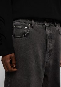 Vaqueros de mezclilla gris con un acabado texturizado, diseño de cinco bolsillos, botón de metal y costuras en contraste. Parte superior negra de manga larga visible.