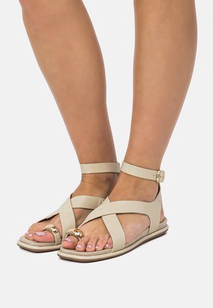 Füße, die beige Sandalen mit offenen Zehen, gekreuzten Knöchelriemen und goldfarbenen Zehenringen auf weißem Hintergrund tragen.