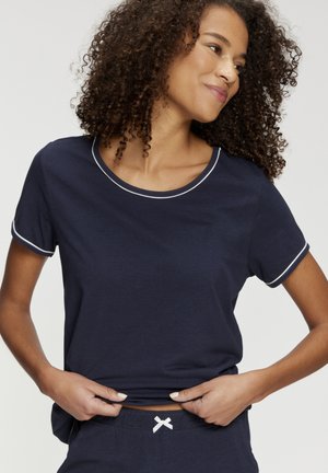 Woman with curly hair wearing navy blue short-sleeve shirt and matching pants, adjusting shirt hem, looking sideways with a slight smile.