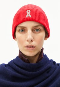 Rode geribbelde gebreide beanie met een wit embleem aan de voorkant. Het model draagt een gestructureerde donkerblauwe sjaal eronder, wat een gelaagde uitstraling creëert.
