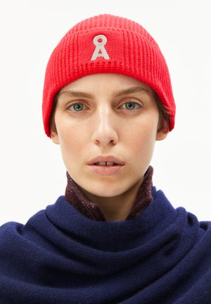 Rode geribbelde gebreide beanie met een wit embleem aan de voorkant. Het model draagt een gestructureerde donkerblauwe sjaal eronder, wat een gelaagde uitstraling creëert.