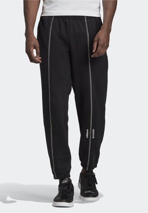 Man draagt zwarte Adidas joggingbroek met witte stiksels en logo's, gecombineerd met zwarte sneakers en een wit shirt, staand tegen een witte achtergrond.