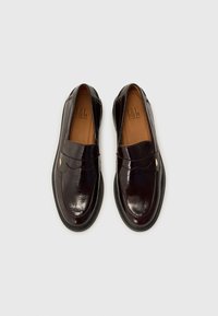 Dunkelbraune Lederloafers mit glänzender Oberfläche, ausgestattet mit einem gewellten Riemen und einem kleinen goldenen Logo-Akzent an der Seite.