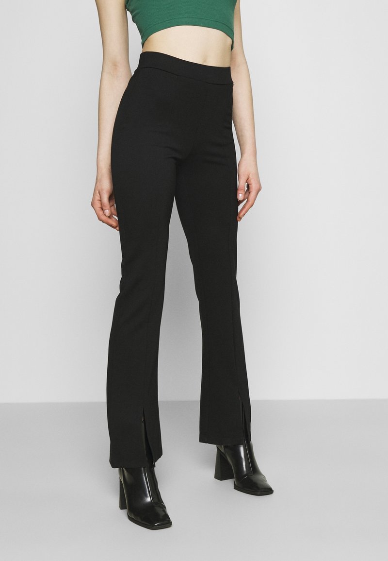 Schwarze Schlaghosen aus glattem Stoff mit vorderen Schlitzen an den Knöcheln und einem hohen, elastischen Bund, kombiniert mit schwarzen Stiefeln mit Absatz.