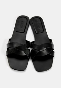 Schwarze flache Sandalen mit einem gekreuzten Riemen-Design. Hergestellt aus glattem Leder mit einer quadratischen Zehenform und einer minimalistischen Sohle.