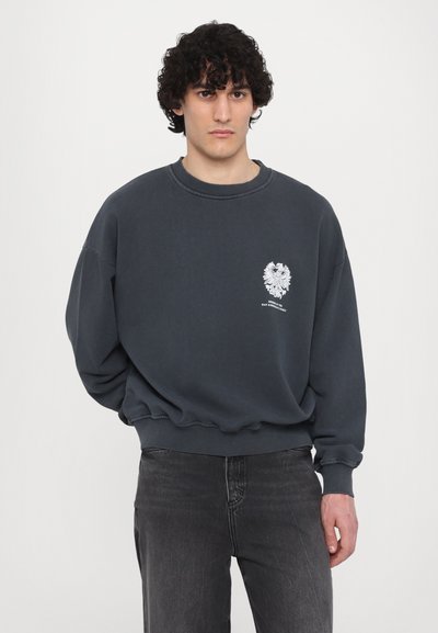 Sudadera gris oscuro hecha de algodón, con un cuello redondo y puños acanalados, y un gráfico de un águila blanca en el área del pecho izquierdo.