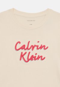 Krémové tričko s krátkým rukávem, které má výrazné vyšívané logo "Calvin Klein" v červené a stříbrné barvě přes hruď. Měkký bavlněný materiál.