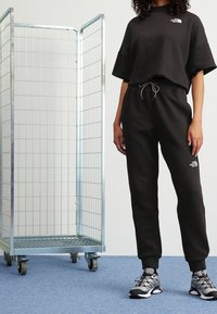 Crop top en maille noire et pantalon de survêtement assorti avec ceinture élastique. Logo argenté sur les deux pièces. Rack à rouleaux en métal en arrière-plan.
