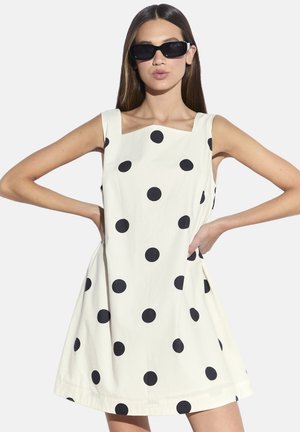 Vestito bianco con grandi pois neri, scollatura quadrata, senza maniche e silhouette svasata. Modella con occhiali da sole neri.
