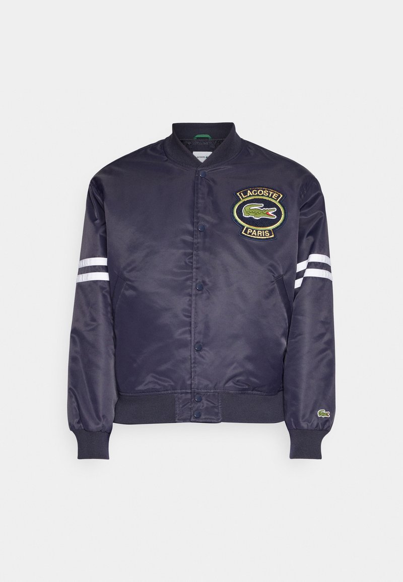 Veste bomber en satin navy avec des poignets et un ourlet côtelés. Présente un logo Lacoste Paris brodé et deux bandes blanches sur les manches.