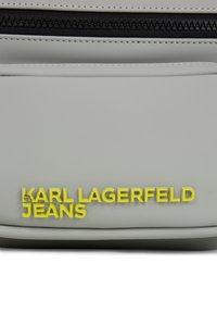 Geantă gri deschis, cu un material texturat și un fermoar negru. Textul galben al logo-ului "KARL LAGERFELD JEANS" este afișat proeminent pe față.