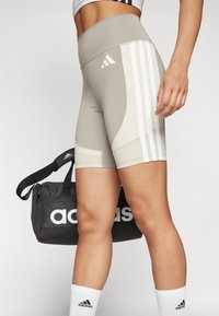 Kerged beežid kõrge vöökohaga sportlikud lühikesed püksid valgete aksenttriipudega ja siledast tekstuurist. Must kott, millel on valge "adidas" logo.