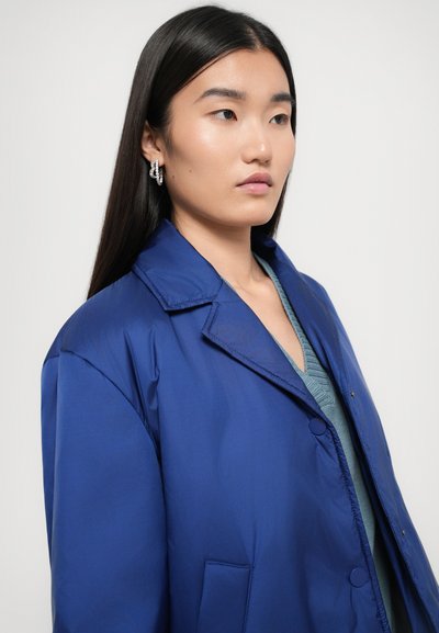 Blå, lett jakke med krage, knappelukking og sidelommer, laget av glatt stoff. Brukt over en strukturert, grønn topp.