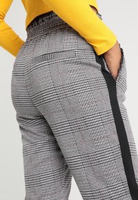 Femme portant un pantalon à carreaux noirs et blancs avec des bandes latérales noires et un haut à manches longues jaune côtelé, la main dans la poche, de dos.