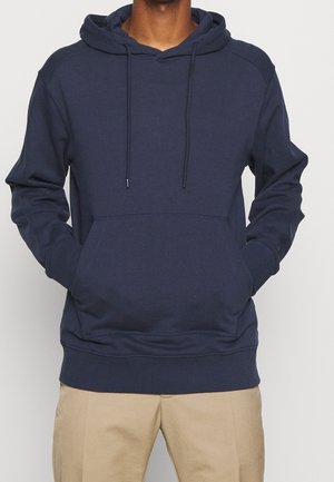 Man in een marineblauwe hoodie met een voorzak en trekkoorden, gecombineerd met beige broek, met de handen in de zakken.