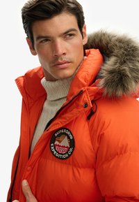 Orange pufferjakke med hue i pels og logo patch. Har en lynlåslukning foran og en striktrøje under. Struktureret, quiltet overflade.