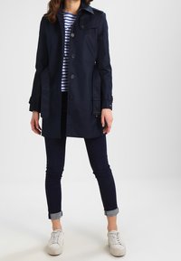 Marinblå trenchcoat med knappstängning, bältesdetalj och sidofickor, lager-på-lager över en randig skjorta, kombinerad med mörka jeans och vita sneakers.