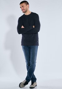 Mann mit verschlossenen Armen, der einen dunklen Strickpullover, blaue Jeans und hellgraue Sneakers mit schwarzen Schnürsenkeln trägt, vor einem schlichten hellen Hintergrund.