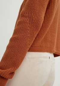 rostfarbener Strickpullover mit strukturiertem Design, ausgestattet mit einem gerippten Saum und Ärmelbündchen, kombiniert mit hellbeigen Cordhosen.