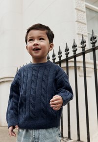 Marineblauwe gebreide trui met kabelpatroon, ronde hals en geribbelde manchetten, gecombineerd met lichtblauwe spijkerbroek. Kind glimlacht en loopt.