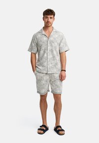 Kurzärmliges, kragenloses, hellgraues Hemd und passende Shorts mit einem strukturierten Muster; getragen mit dunklen Sandalen. Lässiges Sommeroutfit.