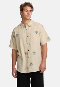 Camicia a maniche corte di colore beige chiaro realizzata in tessuto intrecciato, con motivi circolari verdi e viola, chiusura con bottoni e una singola tasca sul petto.