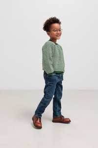 Un pull en tricot rayé vert et blanc, un jean bleu et des chaussures en cuir marron. L'enfant se tient avec les mains dans les poches, souriant et portant des lunettes.