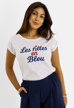 Joven mujer con cabello largo y oscuro lleva una camiseta blanca con el texto "Les filles en Bleu" y pantalones azul marino, de pie con la mano en el bolsillo.