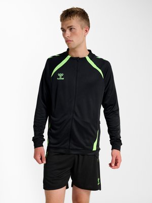 Ung mand iført sort sportjakke med neon grønne accenter og matchende sorte shorts, står og kigger til siden mod en hvid baggrund.