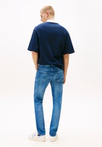 Férfi áll háttal, sötétkék rövid ujjú inget, világoskék farmert és fehér sportcipőt visel fehér háttér előtt.