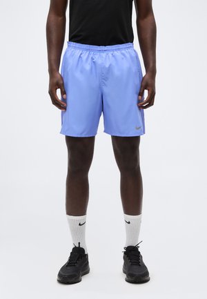 Uomo che indossa pantaloncini sportivi azzurri chiari, scarpe da ginnastica nere, calzini bianchi Nike e una maglietta nera, in piedi davanti a uno sfondo chiaro e uniforme.