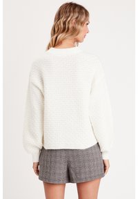 Pull blanc texturé avec un col rond, des manches ballons et des finitions côtelées, associé à un short à carreaux gris.