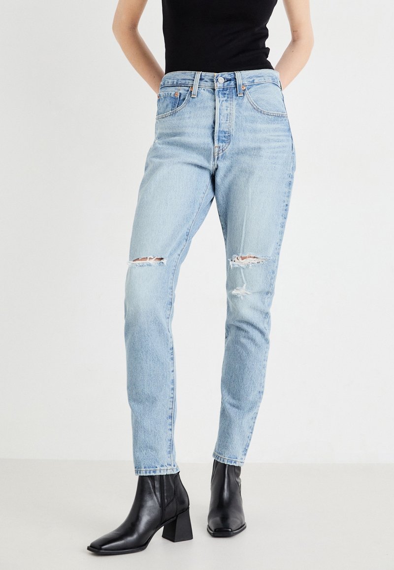 Femme portant un jean skinny déchiré bleu clair et des bottines noires, debout les mains derrière le dos sur fond blanc.
