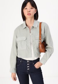 Lys grønn kordfløyelskjorte med knappelukking og to brystlommer, kombinert med mørkeblå high-waisted jeans og en brun crossover-bag.