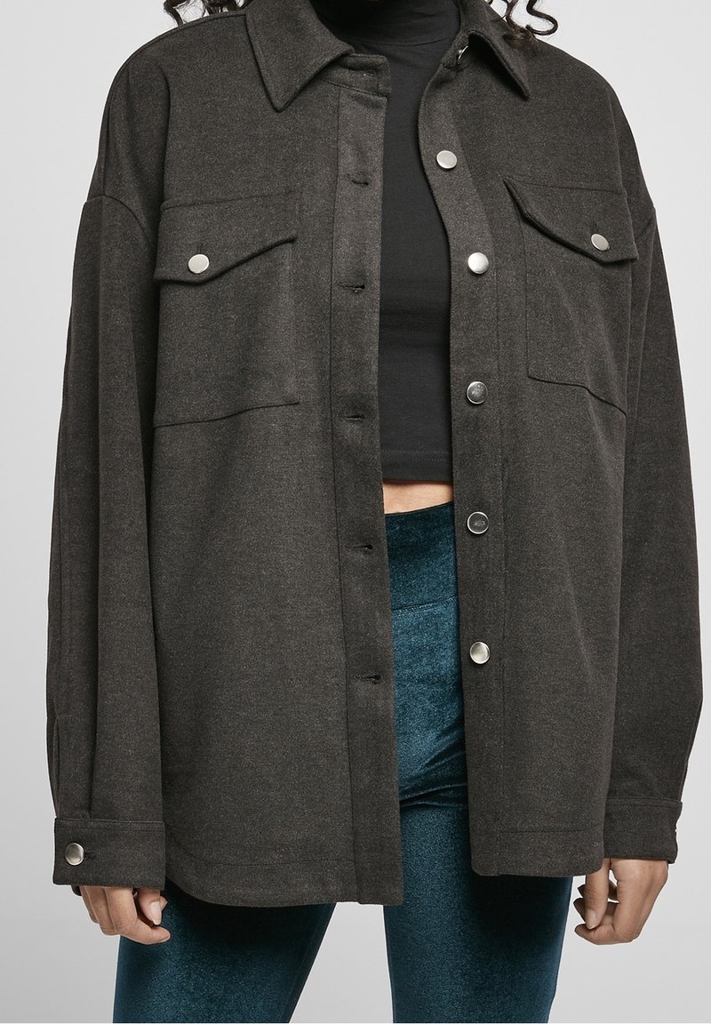 Femme portant une veste oversize gris foncé boutonnée par-dessus un crop top noir et un pantalon en velours bleu, visible des épaules aux hanches.