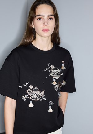 Mujer joven con cabello largo y castaño, vistiendo una camiseta negra de gran tamaño con bordados de teteras, flores y flecos en la parte frontal.