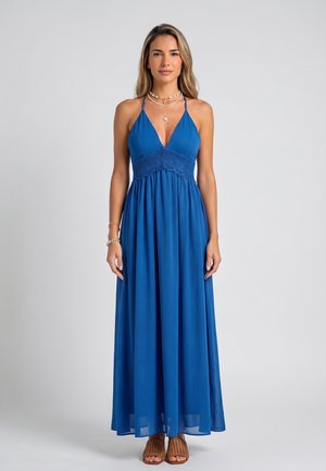 Donna in piedi rivolta in avanti indossa un abito lungo blu senza maniche con dettaglio in pizzo, collane a strati, braccialetti e sandali marroni.