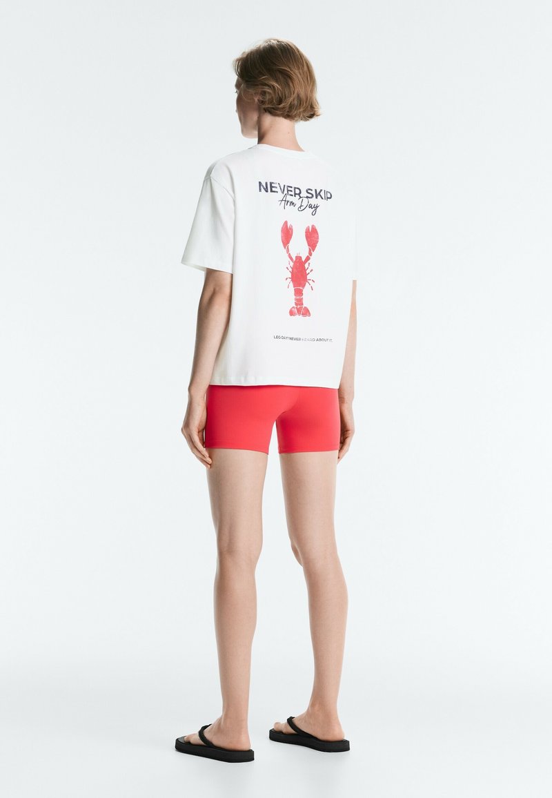 Personne debout, dos tourné, portant un t-shirt blanc avec un homard rouge et le texte "Never Skip Arm Day", et un short rouge avec des sandales noires.